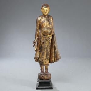 Stående tiggermunk af forgyldt og bemalet teaktræ med krukke i hånden, udskåret dragtbort prydet med farvede glassten. Burma, 19 årh. H. ca. 120 cm.