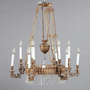 Lysekrone af guldbronzeret metal, behængt med prismer, seks udvendige lysarme. Ca. 1900. H. 92. Diam. 80. Medfølgende lille prismeloftlampe. 2