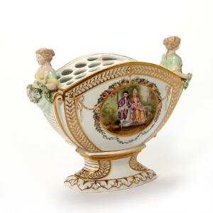 Jardiniere af porcelæn, dekoreret i farver og guld med musikredskaber og figurer i have. Royal Copenhagen. H. 16 cm. L. 23 cm.