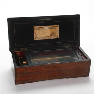 Schweizisk spillemaskine i kasse af palisander dekoreret med intarsia indvendig med mærkat Fabrique De Geneve. 19. årh.s slutning. H. 14 cm. L. 50 cm. D. 23 cm.