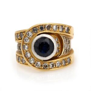 Safir- og diamantring af 18 kt. guld prydet med facetslebet safir omkranset af talrige brillantslebne diamanter. Str. 46,5. Vægt 14 gr. Ca. 1980.