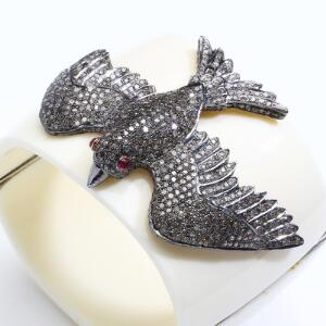 Rubin- og diamantarmring af hvid bakelit med sølv og guld ornamentik i form af fugl prydet med to cabochonslebne rubiner og talrige enkeltslebne diamanter.