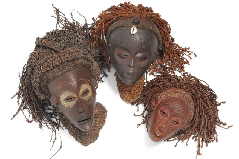 Three masks of carved patinated wood mounted with textiles and raffia with...